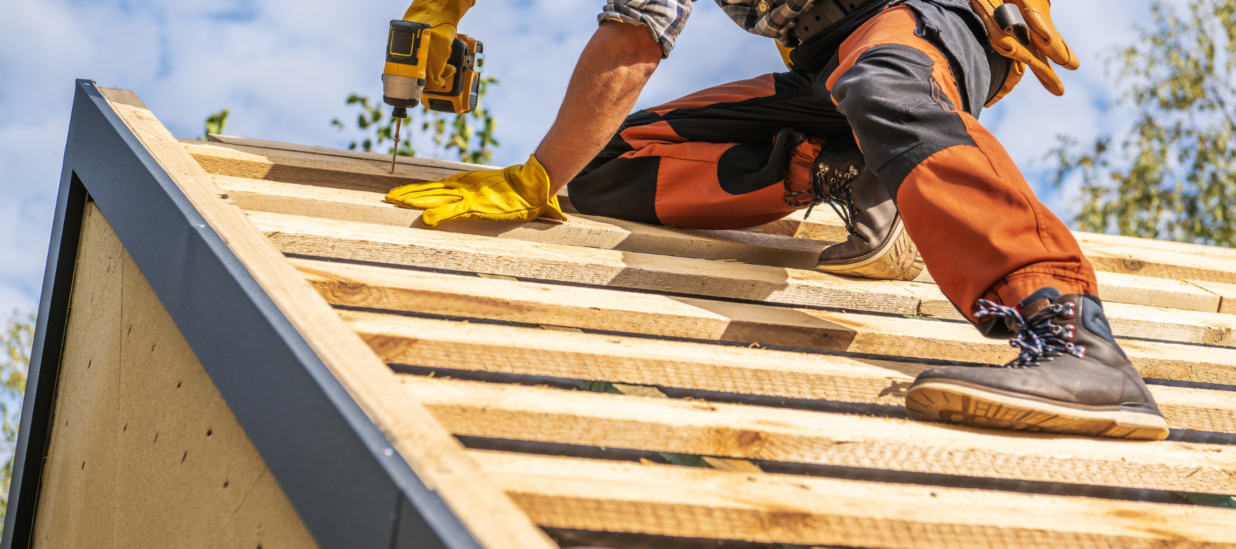 A worker in safety gear installs wooden planks on a roof under a clear sky. The scene conveys focus and diligence in construction work.